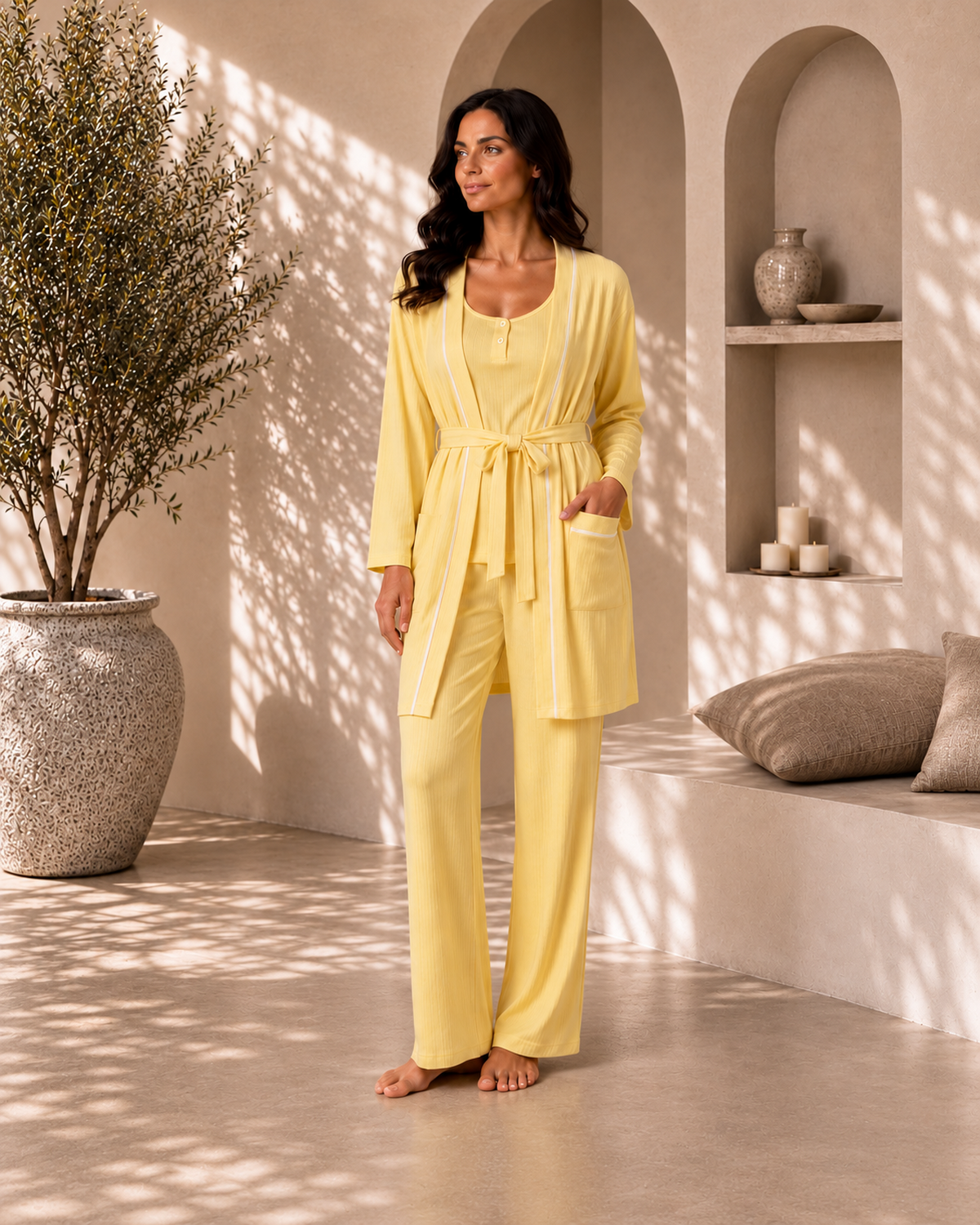 A woman stands barefoot in a sunlit architectural setting wearing a pale yellow matching robe and pants set with white piping details. The scene includes a large textured planter, arched alcoves, candles, and cushions.