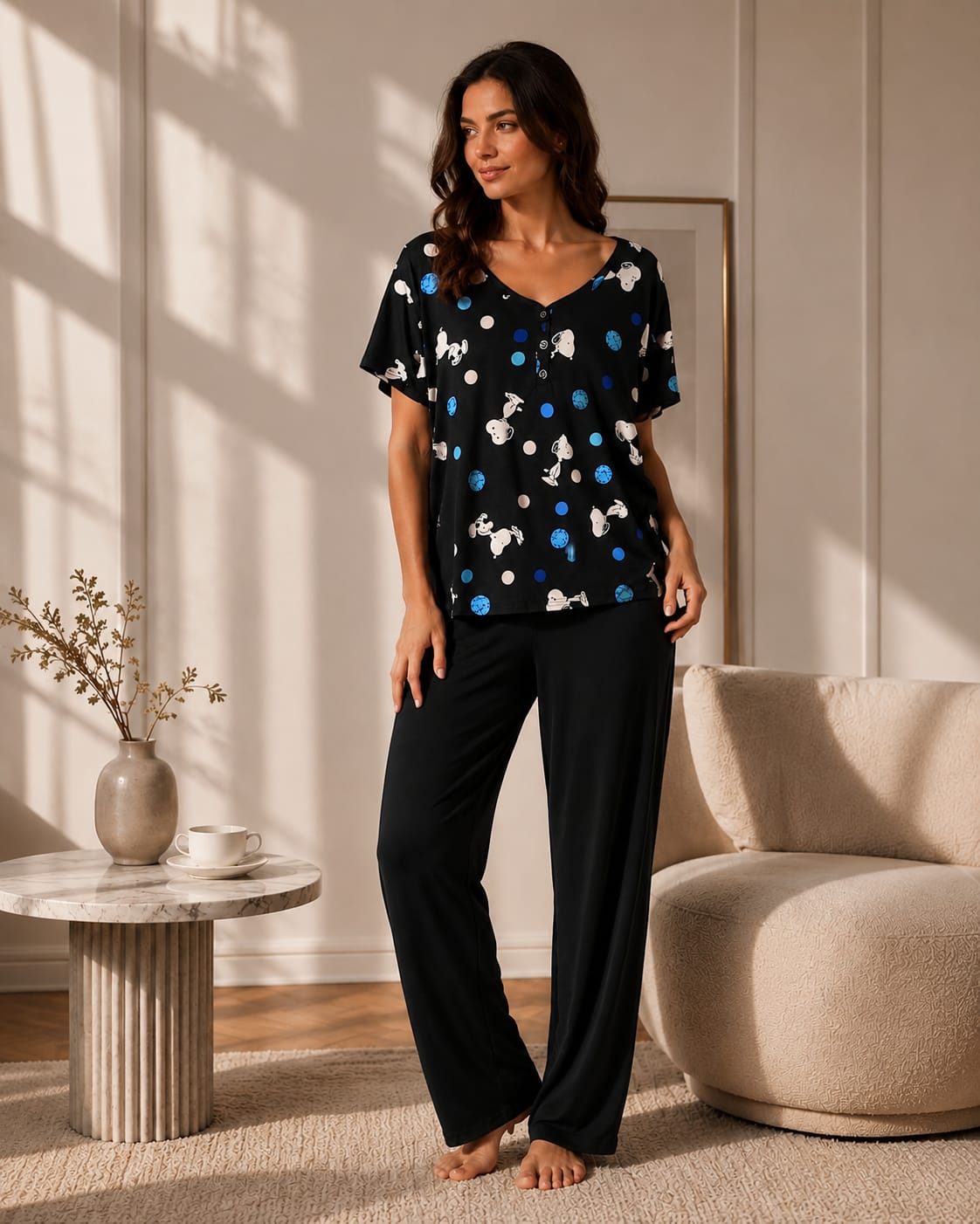 A woman stands barefoot in a sunlit living room wearing a black pajama set with a blue and white dog print on the top and solid black pants.