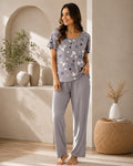 A woman modeling a two-piece pajama set in a warm, modern interior. The set consists of a short-sleeve top with a gray base and a playful print of cartoon animals in blue, white, and black, paired with matching gray pants.