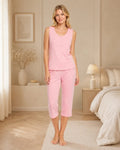 A woman stands barefoot in a softly lit bedroom wearing a light pink sleeveless top and matching pink cropped pants.