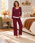 A woman stands barefoot in a bedroom wearing a matching long-sleeve pajama set in burgundy with a white floral print and a white waistband. The set includes a long-sleeve top and wide-leg pants.