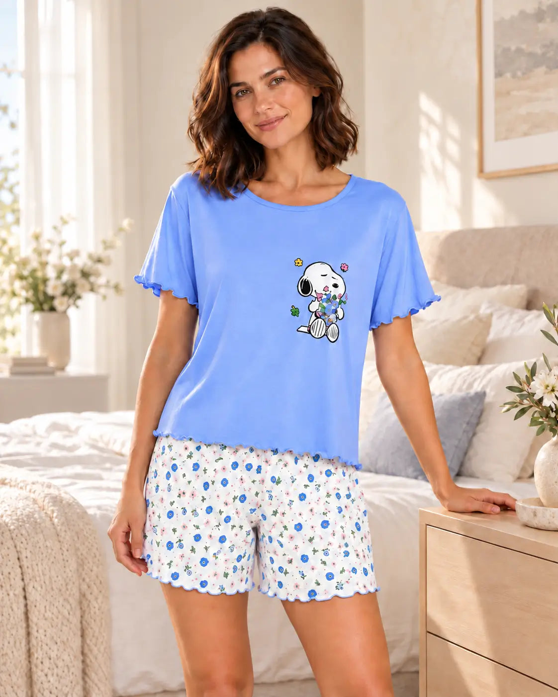 A woman modeling a light blue short-sleeve pajama top with a Snoopy graphic and matching white patterned pajama shorts in a bright bedroom setting.