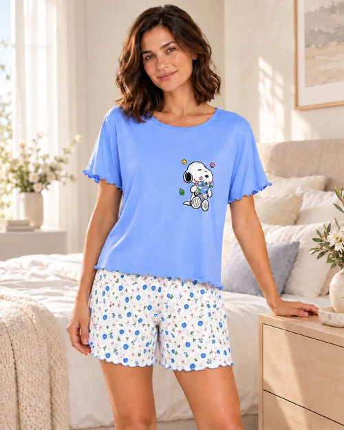 A woman modeling a light blue short-sleeve pajama top with a Snoopy graphic and matching white patterned pajama shorts in a bright bedroom setting.