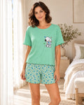 A woman modeling a mint green short-sleeve pajama top with a Snoopy graphic and matching floral pajama shorts in a bright bedroom setting.